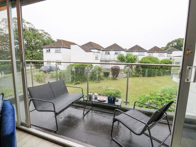 A balcony with a metal bench, chair, and table with tea set overlooking a garden and white houses at 28 Coedrath Park in Saundersfoot