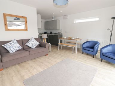 A living room with a brown sofa and two blue chairs next to a dining table with benches and a kitchen area at 26 Coedrath Park in Saundersfoot