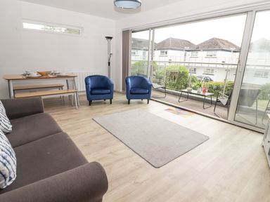 A living room with a sofa, two blue armchairs, a dining table with benches, a floor lamp, and large sliding glass doors leading to a balcony at 26 Coedrath Park in Saundersfoot