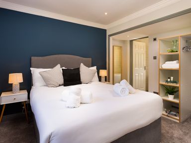 A bedroom with a bed with pillows and rolled towels bedside tables with lamps and shelves with towels and decorative items at Bellman Cottage in Bowness-On-Windermere