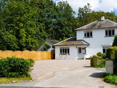 A white two story house with a gravel driveway and wooden fence surrounded by trees at Bellman Cottage in Bowness-On-Windermere