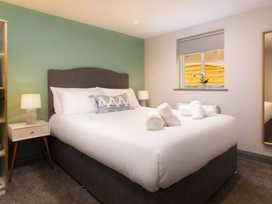 A bedroom with a large bed with white bedding and rolled towels on it two bedside tables with lamps and a small plant on the window sill at Bellman Cottage in Bowness-On-Windermere