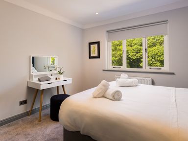 A bedroom with a bed, rolled towels on the bed, a dressing table with a mirror, a stool, and a window showing green trees at Bellman Cottage in Bowness-On-Windermere