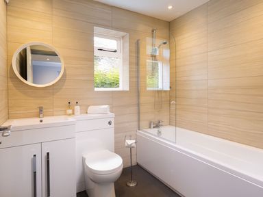 A bathroom with a bathtub and shower combination a toilet a sink with a cabinet round mirror and a small window at Bellman Cottage in Bowness-On-Windermere