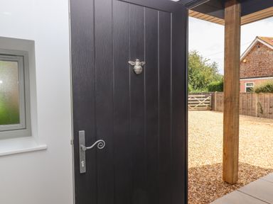 A black exterior door with a silver handle and bee decoration partially open to a gravel yard with a wooden post and fence at Bumblebee Cottage in East Nynehead near Bradford-On-Tone