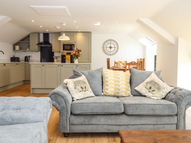 A living room with a grey sofa and animal print cushions next to an open kitchen with light grey cabinets and a dining table at Bumblebee Cottage in East Nynehead near Bradford-On-Tone