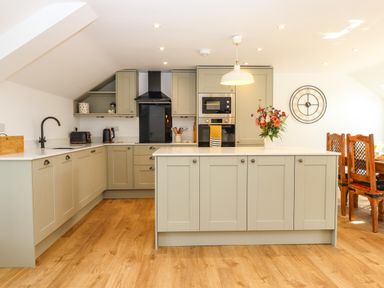 A kitchen with light gray cabinets an island with a vase of flowers wooden chairs and a clock on the wall at Bumblebee Cottage in East Nynehead near Bradford-On-Tone