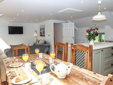 A dining area with a wooden table set with plates of croissants, glasses of orange juice, a teapot, and mugs along with a kitchen island with flowers and a living room with a sofa and wall-mounted television at Bumblebee Cottage in East Nynehead near Bradford-On-Tone