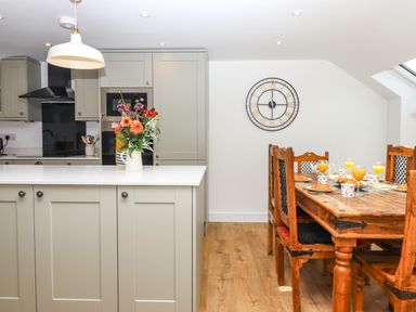 A kitchen with grey cabinets a white countertop and a wooden dining table set with croissants and orange juice at Bumblebee Cottage East Nynehead near Bradford-On-Tone