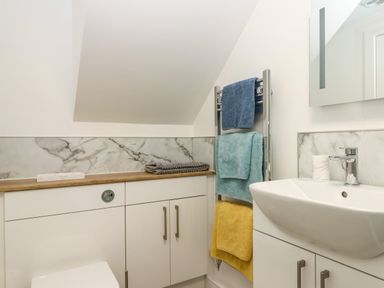 A bathroom with a white sink, marble backsplash, several towels on a towel rack, and white cabinets under a sloped ceiling at Bumblebee Cottage in East Nynehead near Bradford-On-Tone