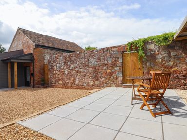 An outdoor patio area with a wooden table and chair set next to a stone wall and a brick building at Bumblebee Cottage in East Nynehead near Bradford-On-Tone