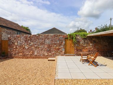 An outdoor patio area with a wooden table and chairs on tiled floor next to a stone wall with a wooden door at Bumblebee Cottage in East Nynehead near Bradford-On-Tone