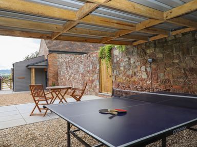 An outdoor covered area with a ping pong table a wooden table and chairs and a stone wall with a wooden door at Bumblebee Cottage in East Nynehead near Bradford-On-Tone