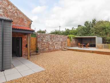 An outdoor courtyard with gravel ground a stone wall wooden gates a covered seating area with chairs and table at Bumblebee Cottage in East Nynehead near Bradford-On-Tone