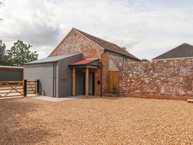 A brick and wood building with a gravel area and a stone wall at Bumblebee Cottage in East Nynehead near Bradford-On-Tone