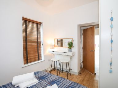 A bedroom with a bed folded towels a window with wooden blinds a wall-mounted desk stools a mirror a lamp and an open door at Gwel Y Mor in Pwllheli