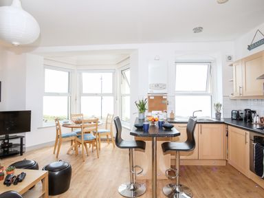 A kitchen and dining area with wooden floor a round dining table by windows and a kitchen island with bar stools at Gwel Y Mor in Pwllheli