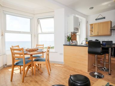 A dining area with a wooden table set for four near large windows and a kitchen with bar stools at Gwel Y Mor in Pwllheli