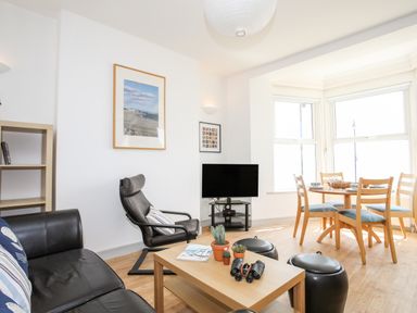 A living room with a black leather sofa a black armchair a wooden coffee table with plants and binoculars and a dining table with wooden chairs at Gwel Y Mor in Pwllheli