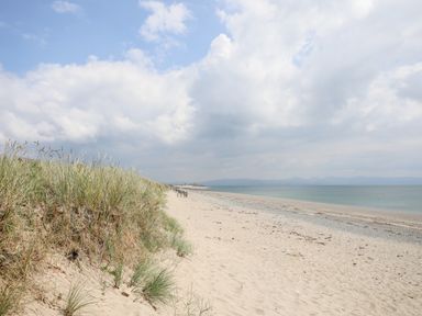 A sandy beach with grassy dunes on the left and the sea on the right under a cloudy sky at Gwel Y Mor in Pwllheli