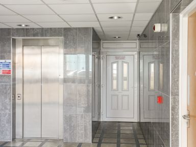 A hallway with an elevator on the left and a white security door at the end with tiled walls and floor at Gwel Y Mor in Pwllheli