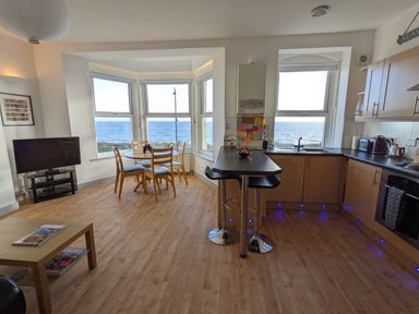 An open kitchen and dining area with a round wooden table and chairs next to large windows overlooking the ocean at Gwel Y Mor in Pwllheli