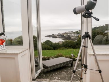 A telescope on a tripod looking out through open doors to a patio with a lounge chair and a coastal village view at Penmarth House in Coverack