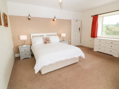 A bedroom with a double bed white bedding bedside tables lamps a chest of drawers and a window with red curtains at Penmarth House in Coverack