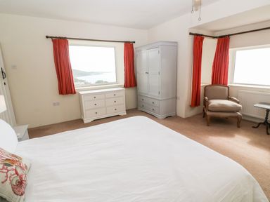 A bedroom with a bed dresser armoire armchair and windows with red curtains at Penmarth House in Coverack