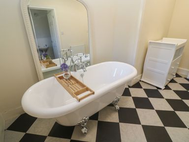 A bathroom with a clawfoot bathtub a wooden bath tray with flowers and a glass on it a large mirror and a white storage cabinet at Penmarth House in Coverack