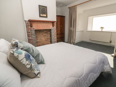 A bedroom with a bed, pillows, a brick fireplace, a wooden cabinet, a window with curtains, and a vase on the windowsill at Penmarth House in Coverack