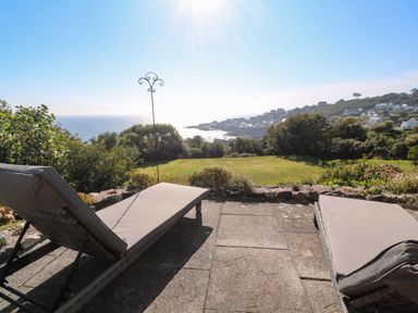A patio with two lounge chairs overlooking a grassy lawn and ocean with houses in the distance at Penmarth House in Coverack