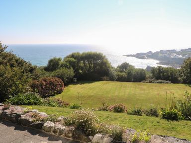 A garden with grass lawn, bushes, and trees overlooking the sea and a coastal village at Penmarth House in Coverack