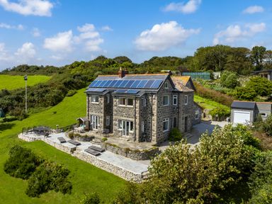 A two-story stone house with solar panels on the roof and a stone patio with outdoor furniture in a green rural area at Penmarth House in Coverack