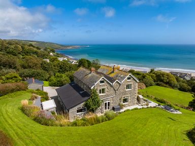 A stone house with a black roof surrounded by green lawns and trees near the coast with a beach and ocean in the background at Penmarth House in Coverack