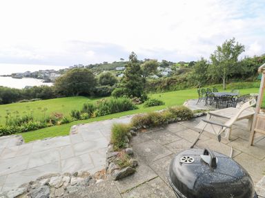 A garden with patio furniture and a barbecue overlooking a grassy lawn and coastal houses at Penmarth House in Coverack