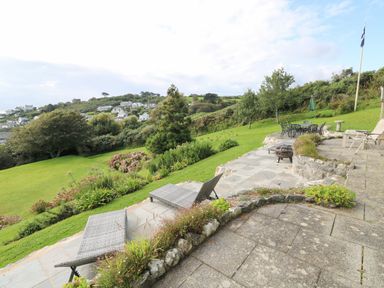 An outdoor patio area with lounge chairs and a dining table set on a tiled surface overlooking a grassy garden with trees and bushes at Penmarth House in Coverack