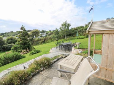 An outdoor patio area with a table and chairs next to a wooden structure and grassy garden at Penmarth House in Coverack