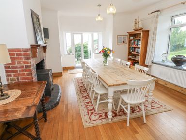 A dining room with a wooden table and white chairs a red patterned rug a wooden bookshelf a fireplace and glass doors at Penmarth House in Coverack