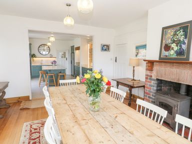 A dining room with a wooden table and white chairs a vase of flowers a fireplace and a kitchen in the background at Penmarth House Coverack