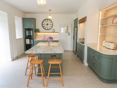 A kitchen with green cabinets a large island with stools and a wall clock at Penmarth House in Coverack