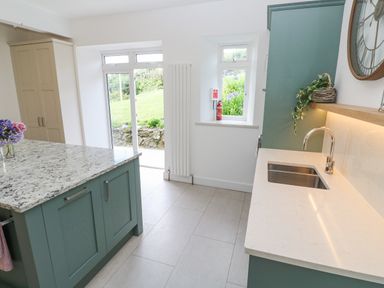 A kitchen with green cabinets a marble island countertop a sink a window a door and a clock on the wall at Penmarth House in Coverack