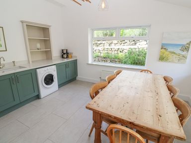 A kitchen with a washing machine green cabinets a wooden dining table with chairs and a large window showing a stone wall garden at Penmarth House in Coverack