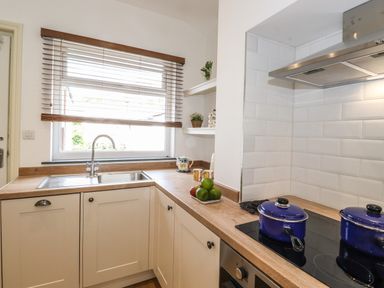 A kitchen with a sink under a window with wooden blinds pots on a stove and shelves with decorations at Halewood in Bowness-On-Windermere