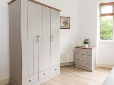 A bedroom corner with a wardrobe and chest of drawers with a plant on top and a window showing greenery at Halewood in Bowness-On-Windermere