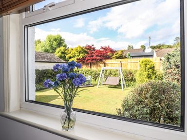 A vase with blue flowers on a windowsill looking out to a garden with a swing bench and various trees and bushes at Halewood in Bowness-On-Windermere