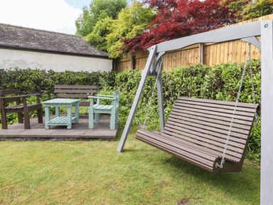 A garden with a wooden swing and outdoor chairs and table on a paved area at Halewood in Bowness-On-Windermere