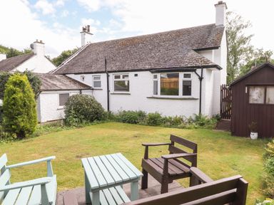 A garden with wooden chairs and table in front of a white house with a tiled roof and a brown shed at Halewood in Bowness-On-Windermere