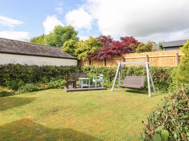 A garden with a wooden swing bench a wooden bench and two chairs around a small table surrounded by hedges at Halewood in Bowness-On-Windermere