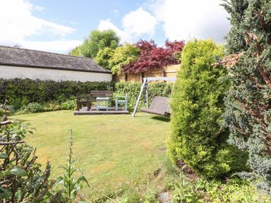 A garden with wooden chairs and a table on a deck and a wooden swing surrounded by trees and bushes at Halewood in Bowness-On-Windermere